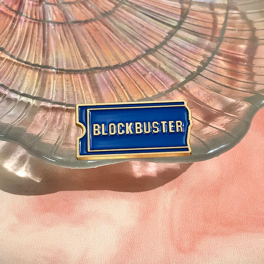 A rectangular enamel pin with the word 'BLOCKBUSTER' in bold blue letters on a gold-colored background, placed on a surface with a reflective background and a pink-ish tint.