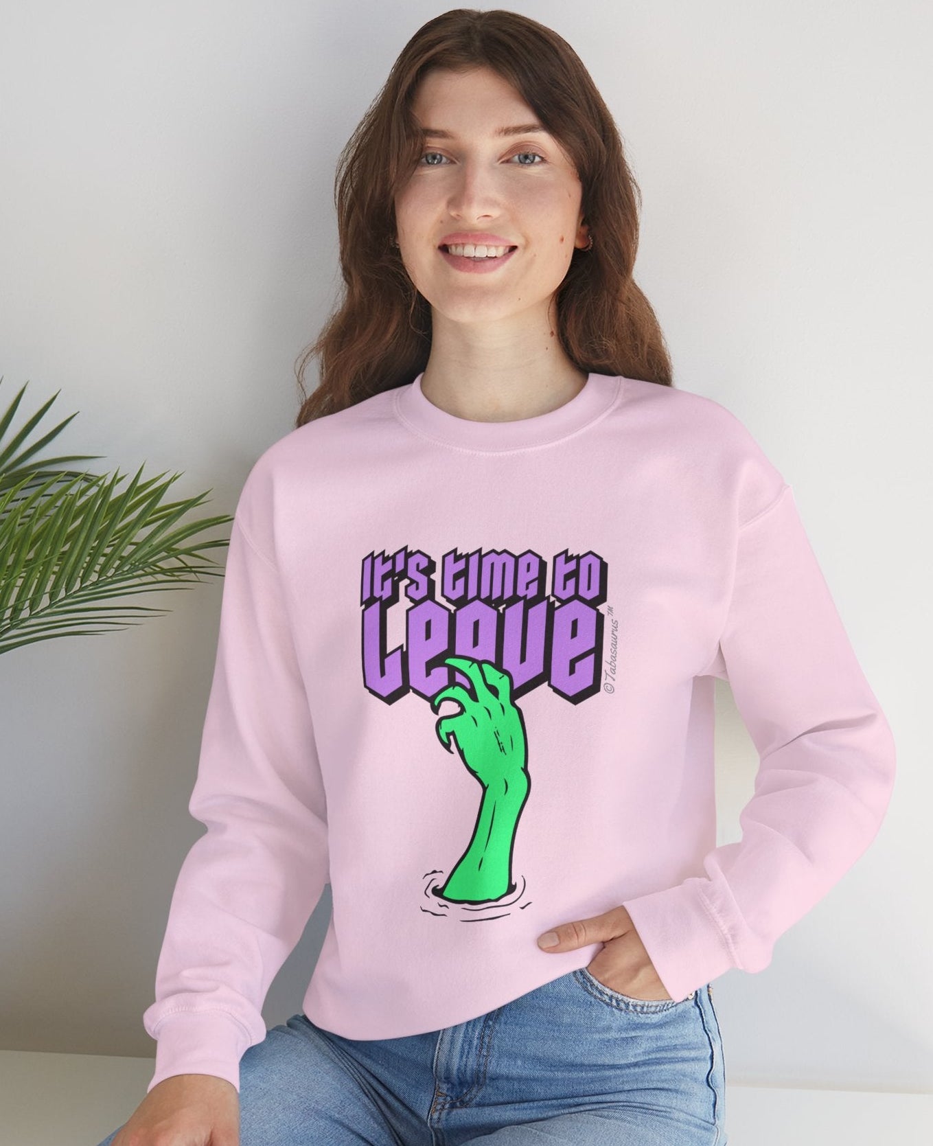 Person wearing a pink sweatshirt with a graphic design and text, sitting on a white surface with a plant in the background.